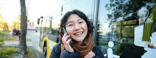 cellulair technologie en mensen concept. elegant Aziatisch meisje praat Aan mobiel telefoon, maakt een telefoon telefoongesprek, staat in de buurt bus hou op en heeft gesprek foto
