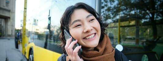 dichtbij omhoog portret van gelukkig Aziatisch meisje praat Aan mobiel telefoon, glimlacht terwijl spreekt naar iemand Aan smartphone, staat in de buurt bus Aan hou op foto
