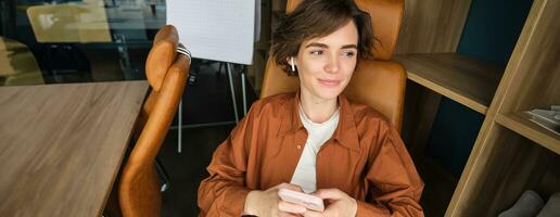 portret van vrouw werken in een kantoor, werknemer gebruik makend van smartphone, zittend in stoel Aan haar breken en glimlachen foto