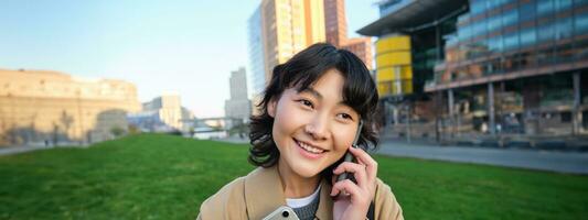 Universiteit levensstijl. jong Aziatisch leerling, meisje met tablet en notitieboekje, praat Aan mobiel telefoon terwijl wandelingen van campus Aan straat foto