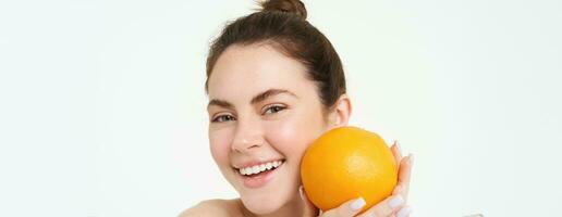 vitamine c en Gezondheid concept. jong schoonheid vrouw, Holding oranje fruit in de buurt gezicht, concept van biologisch huidsverzorging behandeling, gelaats en lichaam producten, staand over- wit achtergrond foto