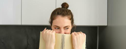 schattig vrouw verbergt gezicht achter notitieboekje, op zoek terzijde, glimlachen met ogen foto