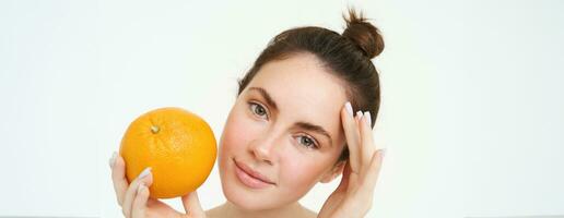 dichtbij omhoog portret van brunette vrouw model, Holding oranje in de buurt gezicht, staand over- wit achtergrond foto