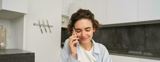 portret van mooi meisje praat Aan mobiel telefoon, heeft gesprek, antwoord een telefoontje in keuken foto