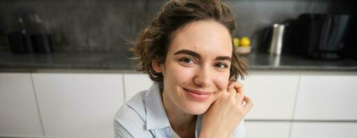 dichtbij omhoog portret van jong brunette vrouw, looks Bij camera en glimlacht, zit Bij huis in keuken foto