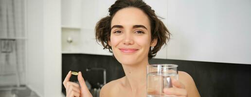 dieet supplementen en gezond levensstijl. jong vrouw nemen vitamine c, d omega-3 met glas van water, staand in actieve kleding, drinken na training opleiding in haar keuken foto