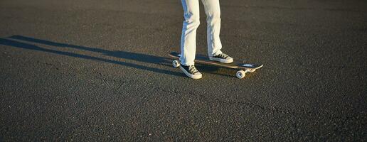 bijgesneden schot van poten Aan longboard. schaatser meisje rijden haar skateboard Aan straat. vrouw tiener Aan kruiser foto