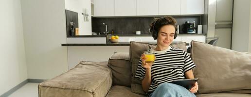 portret van vrouw in koptelefoon, zittend Aan sofa met tablet en drinken oranje sap, aan het kijken TV tonen Aan haar apparaatje, gebruik makend van sociaal media sollicitatie, uitgeven tijd Bij huis foto