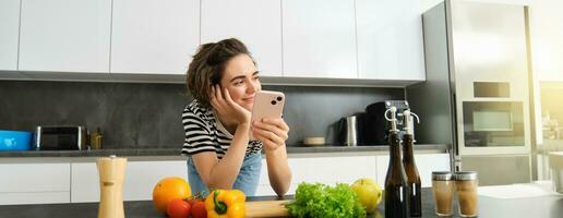 portret van jong glimlachen vrouw, zoeken Koken recept online Aan smartphone, staand in de buurt groenten en hakken bord, maken maaltijd, voorbereidingen treffen salade en gebruik makend van sociaal media Aan mobiel telefoon foto