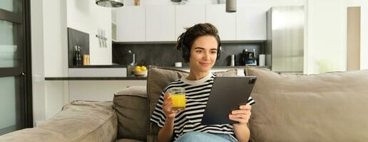 portret van vrouw in koptelefoon, zittend Aan sofa met tablet en drinken oranje sap, aan het kijken TV tonen Aan haar apparaatje, gebruik makend van sociaal media sollicitatie, uitgeven tijd Bij huis foto