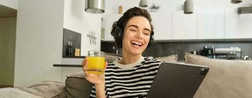 portret van vrouw in koptelefoon, zittend Aan sofa met tablet en drinken oranje sap, aan het kijken TV tonen Aan haar apparaatje, gebruik makend van sociaal media sollicitatie, uitgeven tijd Bij huis foto