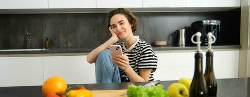 portret van jong vrouw zoeken voor Koken recepten online gebruik makend van smartphone, zittend in de buurt groenten, salade ingrediënten en hakken bord, glimlachen Bij camera foto