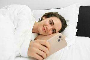 beeld van vrouw model- in bed, op zoek Bij mobiel telefoon, lezing Aan smartphone app, glimlachen verheugd foto