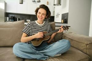jong vrouw spelen ukulele met passie, het zingen liedje, tokkelen snaren, zittend Aan sofa in leven kamer Bij huis foto