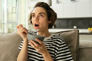 dichtbij omhoog portret van vrouw aan het eten in voorkant van TV, op zoek versteld staan en met interesseren Bij scherm, aan het eten ontbijt, Holding kom van granen en lepel, zittend Aan sofa gefascineerd foto