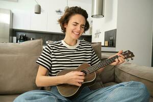 portret van jong modern vrouw, leerling spelen ukulele Bij huis, zittend met klein gitaar, het zingen en gevoel Vrolijk, zittend Aan sofa foto