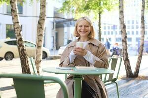 portret van elegant modern vrouw, zittend in een buitenshuis cafe, glimlachen en drinken koffie van meenemen beker, vervelend loopgraaf jas foto