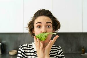 dichtbij omhoog van grappig schattig vrouw, vegetarisch aan het eten sla blad en lachend, concept van gezond eetpatroon, meisje houdt groenten, staand in de keuken foto