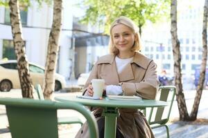 portret van elegant modern vrouw, zittend in een buitenshuis cafe, glimlachen en drinken koffie van meenemen beker, vervelend loopgraaf jas foto