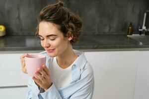 portret van een blij jong vrouw genieten van een kop van koffie Bij huis. glimlachen mooi meisje drankjes heet thee in ochtend. opgewonden vrouw lachend Aan een herfst dag foto