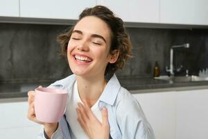 portret van een blij jong vrouw genieten van een kop van koffie Bij huis. glimlachen mooi meisje drankjes heet thee in ochtend. opgewonden vrouw lachend Aan een herfst dag foto