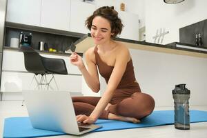 beeld van jong geschiktheid meisje, vrouw atleet Bij huis, training Aan keuken vloer, Holding credit kaart met laptop, inkoop online opleiding sessie, Sportschool toepassing voor huis training foto