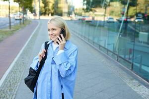 vrolijk jong modern vrouw wandelen Aan straat en antwoorden telefoon telefoongesprek, pratend Aan mobiel, draagt zwart rugzak foto