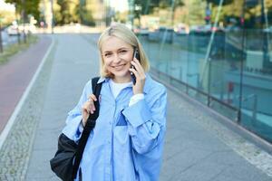 portret van jong glimlachen vrouw pratend Aan mobiel telefoon, leerling wandelingen huis met smartphone, draagt zwart rugzak foto