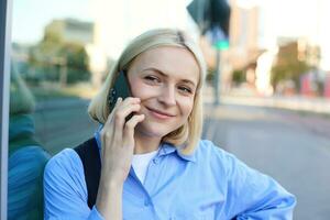 dichtbij omhoog portret van glimlachen blond vrouw, chatten Aan de telefoon, pratend Aan mobiel telefoon, staand Aan straat buitenshuis foto