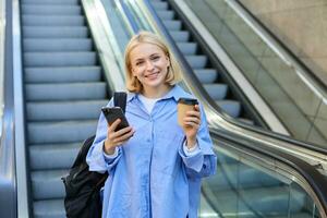 beeld van elegant jong vrouw in blauw shirt, Holding meenemen kop van koffie en mobiel telefoon, staand in de buurt roltrap en aan het wachten voor jij, glimlachen Bij camera foto
