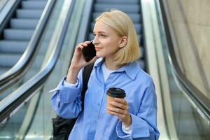 portret van elegant jong vrouw met kop van koffie, staand in de buurt roltrap in stad centrum, antwoord telefoon telefoongesprek, gebruik makend van smartphone, pratend naar iemand foto