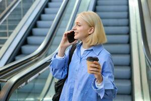 portret van schattig jong blond vrouw, leerling met kop van koffie, antwoorden telefoon telefoongesprek, staand in de buurt roltrap in stad en praten, chatten met iemand over- de smartphone foto