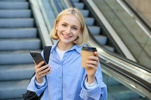 portret van jong mooi vrouw, meisje leerling met kop van koffie en smartphone, staand in de buurt roltrap in stad centrum, glimlachen en op zoek Bij camera foto