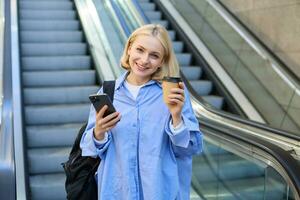 beeld van mooi vrouw model, leerling staand in de buurt roltrap, verhogen kop van meenemen koffie, glimlachen en op zoek Vrolijk, heeft mobiel telefoon in hand- en rugzak Aan schouder foto