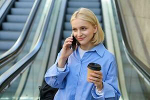 portret van schattig jong blond vrouw, leerling met kop van koffie, antwoorden telefoon telefoongesprek, staand in de buurt roltrap in stad en praten, chatten met iemand over- de smartphone foto