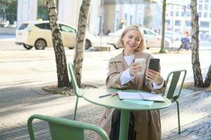 levensstijl portret van jong vrouw heeft video gesprek, chatten Aan smartphone app, tonen meenemen kop terwijl zittend buiten in koffie winkel foto