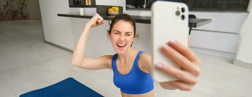 jong energiek meisje doet sport- Bij huis, shows haar spieren Aan arm en duurt selfie Aan smartphone, poseren Aan geschiktheid rubber mat in leven kamer foto