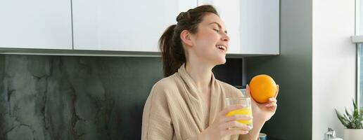 portret van gelukkig vrouw in keuken, vervelend badjas, drinken oranje sap, vers geperst drankje, glimlachen en lachend, voedsel en drinken concept foto