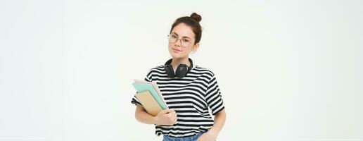 onderwijs concept. glimlachen brunette meisje, leerling in gewoontjes kleren, houdt haar boeken, studie materiaal, draagt hoofdtelefoons over- nek, looks zelfverzekerd en Vrolijk, geïsoleerd over- wit achtergrond foto