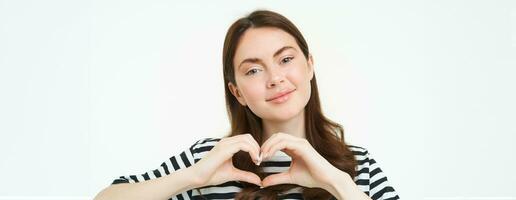 dichtbij omhoog portret van schattig jong vrouw shows hart teken, staren Bij camera met liefde en zorg, staand over- wit achtergrond foto