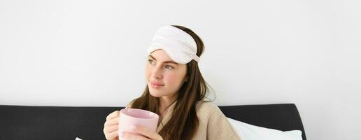 prachtig vrouw in pyjama en slapen masker, Holding kop van koffie, drinken cappuccino in haar bed en op zoek Vrolijk, resting in slaapkamer Aan weekend, hebben ontspannende ochtend- foto