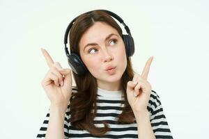 portret van jong brunette vrouw in koptelefoon, luisteren muziek- in koptelefoon, richten vingers omhoog, tonen banier, advertentie, geïsoleerd over- wit achtergrond foto