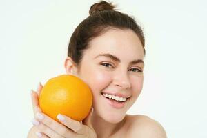 vitamine c en Gezondheid concept. jong schoonheid vrouw, Holding oranje fruit in de buurt gezicht, concept van biologisch huidsverzorging behandeling, gelaats en lichaam producten, staand over- wit achtergrond foto