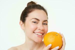 dichtbij omhoog portret van glimlachen jong vrouw, Holding oranje in de buurt gezicht, op zoek Vrolijk, concept van vitamines, biologisch en natuurlijk huidsverzorging behandeling met c serum, wit achtergrond foto