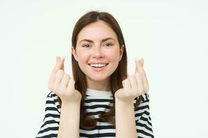 portret van schattig jong brunette vrouw, shows kawaii vinger harten en lachend, houdt iets, staat in gestreept t-shirt over- wit achtergrond foto