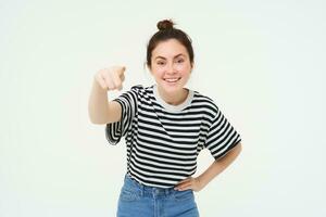 beeld van elegant jong vrouw, richten vinger Bij camera, uitnodigend jij, staand over- wit achtergrond foto