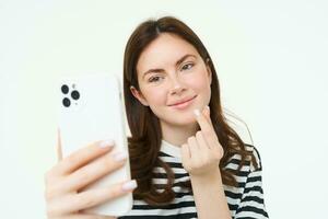 portret van mooi vrouw tonen hart teken en poseren voor selfie, nemen afbeelding van haarzelf Aan smartphone app, poseren in de buurt iets schattig, wit achtergrond foto