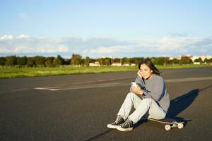 schaatser meisje zit Aan haar skateboard Aan weg, gebruik makend van smartphone, chatten Aan mobiel app foto