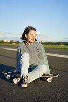 schattig glimlachen Aziatisch meisje zit Aan longboard, skateboarden alleen Aan een leeg weg. kopiëren ruimte foto