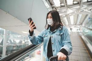 Aziatische vrouwenreiziger die masker draagt die smartphone gebruiken die zich op roltrap bevindt foto